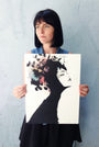 A person with short black hair holds a Conrad Roset limited edition fine art giclée print against a textured gray wall, showcasing a stylized womans head adorned with vibrant flowers and foliage, reminiscent of his style, as they gaze thoughtfully into the distance.