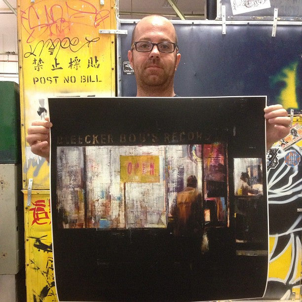 A person holds a large, blurry photo of Bleecker Bobs storefront, featuring vibrant graffiti and posters, capturing the essence of a Brett Amory limited edition fine art giclee.