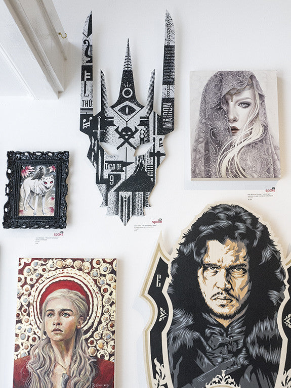 A wall at Spoke Art gallery features diverse Derek Ballard artworks: a wolf portrait in a black frame, an abstract monochrome mask, a veiled woman, a crowned woman, and The Dispensation of Sauron—a striking screen ink on aluminum piece among vivid characters.