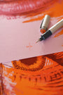 A close-up of a Bartosz Kosowski-style artwork showcases an eye in vivid orange and red tones. In front, a pink sheet marked 24/30 in red ink rests by the screenprint, beside an open black pen with its cap ready for use.