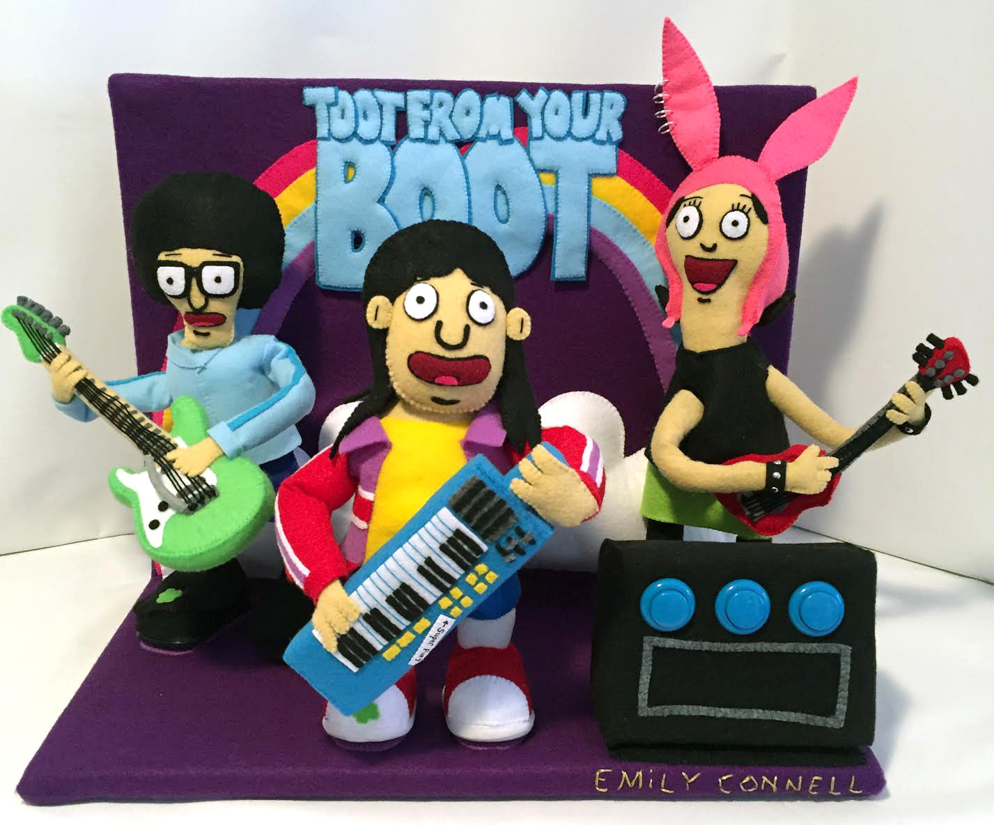 Felt figurines from Emily Connells collection play instruments on a stage with the Toot From Your Boot backdrop. One plays a keyboard, another a green guitar, and the last a red guitar, all crafted with lively felt materials reminiscent of Bobs Burgers art at Spoke Art NYC.