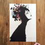 A black silhouette of a woman in profile with floral elements intertwined in her hair, splashed with color against a wooden background, features a hand holding a pen at the bottom right. This piece is reminiscent of fine art giclée prints by Conrad Roset.