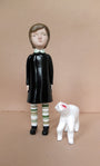 A Doubleparlour ceramic figurine features a child in a black dress with striped leggings, intricately hand sculpted from resin, alongside a small white ceramic dog against a plain beige backdrop.