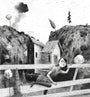 Jonas Lofgren presents a graphite on paper illustration of a person leaning on a wooden fence, gazing into the distance. In the backdrop, nestled among rocky hills, sits a house. This serene black and white scene is complete with trees and clouds in the sky.