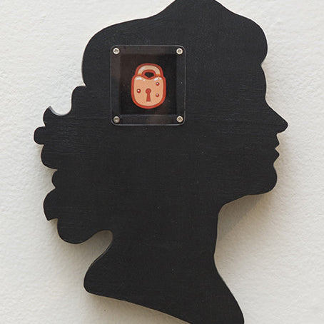 A hand-cut wood silhouette of a human head profile in black by Matt Ritchie is mounted on a light wall, featuring a whimsical red cartoon lock with a face, reminiscent of an art gallery acrylic piece.