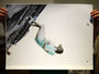 Someone holds a Henrik Uldalen fine art giclee print showing a woman in a light blue dress, floating sideways against an abstract cityscape in gray and black. The piece is part of a signed and numbered edition with an embossed seal on the lower left corner.