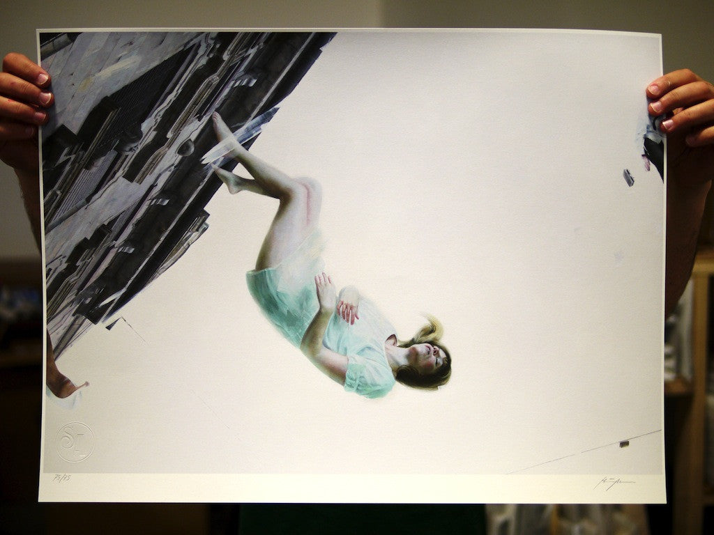Someone holds a Henrik Uldalen fine art giclee print showing a woman in a light blue dress, floating sideways against an abstract cityscape in gray and black. The piece is part of a signed and numbered edition with an embossed seal on the lower left corner.