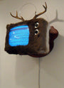 A wall-mounted TV by Jeremiah Jenkins, designed to resemble a taxidermy deer with antlers, a deer hide, and googly eyes. The screen crackles with static.