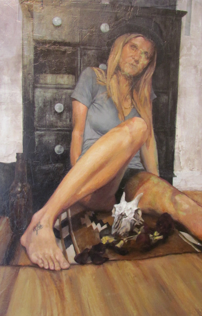 In an oil on wood panel by Elliot Brown, a blonde woman in a gray shirt and hat sits elegantly against a dark dresser. Her tattooed foot is extended amid a skull, roses, and bottle on a patterned rug—creating an artwork suited for any gallery.