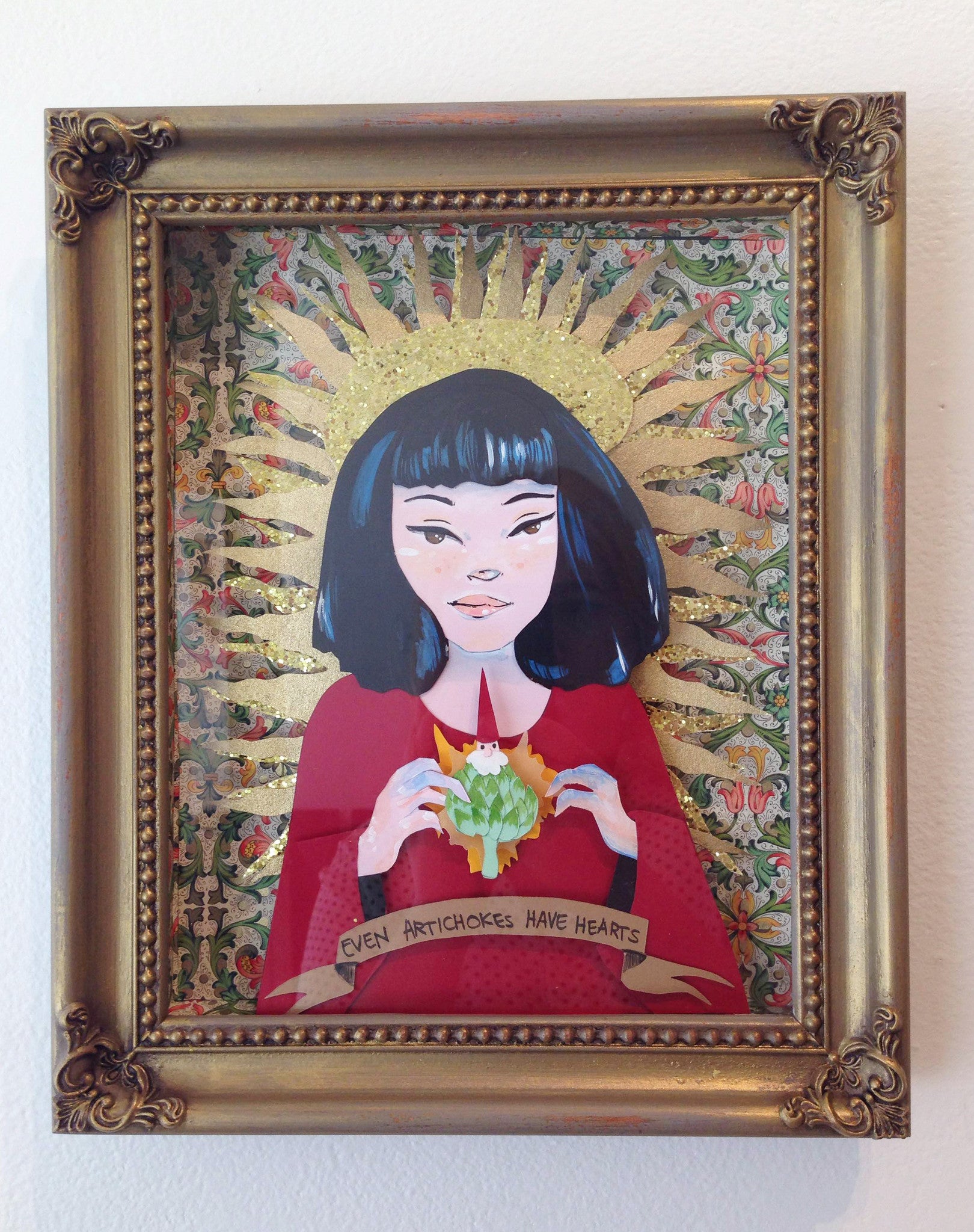 Jill Bencsits presents a framed artwork depicting a black-haired woman holding an artichoke, wearing red against a floral backdrop with a golden halo. The shadow frame features the banner Even Artichokes Have Hearts, and the ornate frame is gold-toned.