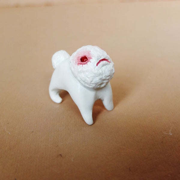 This hand-sculpted resin creation by Doubleparlour portrays Little Bashlu, a small four-legged creature with a textured face and sad expression, highlighted by striking red eyes. It gracefully stands on a light brown surface, hinting at untold stories.