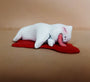 A small resin sculpture by Doubleparlour features a white cat playfully lying on its side with its tongue out, atop a red puddle base. Hand-sculpted and finished in acrylic paint, it embodies whimsical charm against a neutral backdrop.