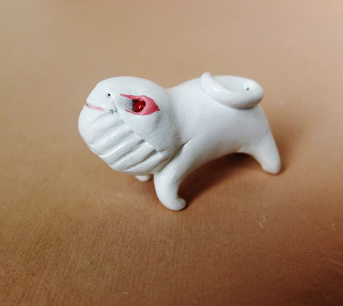 A small, hand-sculpted Doubleparlour pug-like figurine in white resin features exaggerated traits like a wrinkled face and curled tail. Painted with acrylics, it displays vivid red around the eyes, seated on brown, evoking the whimsy of the Little Bashlu series.