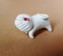 A small, hand-sculpted Doubleparlour pug-like figurine in white resin features exaggerated traits like a wrinkled face and curled tail. Painted with acrylics, it displays vivid red around the eyes, seated on brown, evoking the whimsy of the Little Bashlu series.