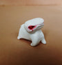 A small white figure with a round body, short legs, and a long head featuring a pink-red eye stands against a brown backdrop. Hand sculpted from resin and painted with acrylics, this exquisite piece is crafted by Doubleparlour.