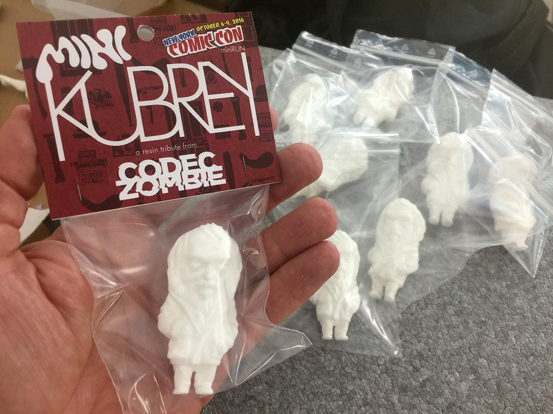 A hand holds a small white figurine in a bag alongside others on a table. The label reads Mini Kubrey by Codeczombie with a Comic-Con nod. This limited edition action figure features an abstract design, making it an exceptional collectible for enthusiasts.