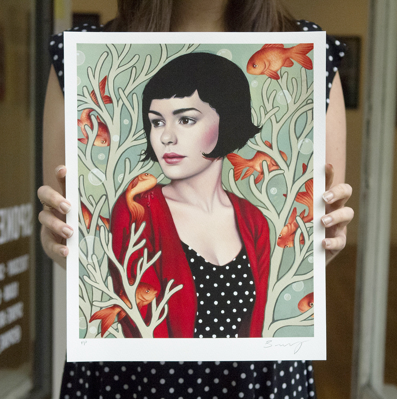 A person holds a Sarah Joncas limited edition giclee, featuring a woman with short black hair in a red cardigan and polka dot dress, surrounded by coral and orange fish on a light green background. Its perfect for Valentines Day delivery.