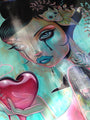 A vibrant illustration titled Stitched Up by Caia Koopman features a stylized woman with large eyes, blue lips, and a tear on her cheek. She holds a stitched red heart, with hair adorned by a skull and leaves, set against an acrylic wood panel with turquoise abstract elements.