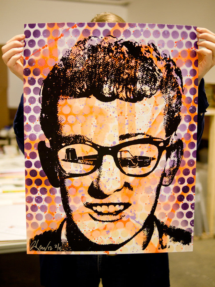 A person displays a pop art poster by Rene Gagnon, reminiscent of Buddy Holly, with a portrait featuring NIKON in glasses. The screen print is signed, numbered, and set against an orange and purple dot backdrop for an authentic touch.