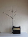A Jeremiah Jenkins black vintage radio is placed on a white surface against a textured white wall, with a mixed-media sculpture resembling tree branches attached to it, creating a minimalist and artistic setup.