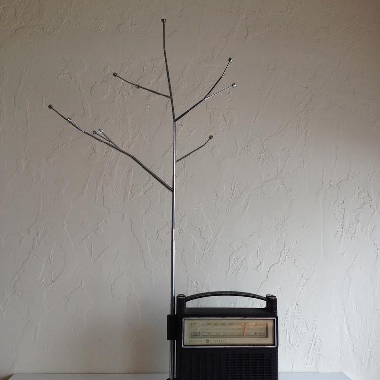 A Jeremiah Jenkins black vintage radio is placed on a white surface against a textured white wall, with a mixed-media sculpture resembling tree branches attached to it, creating a minimalist and artistic setup.