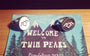 Shaun OKeefe cufflinks with abstract geometric designs are showcased on a postcard with the iconic Welcome to Twin Peaks sign, set against a forest and mountain backdrop, echoing the mystery of a custom Twin Peaks ring.
