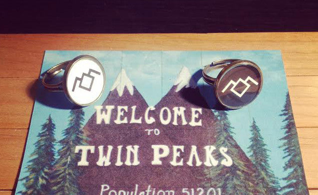 Shaun OKeefe cufflinks with abstract geometric designs are showcased on a postcard with the iconic Welcome to Twin Peaks sign, set against a forest and mountain backdrop, echoing the mystery of a custom Twin Peaks ring.
