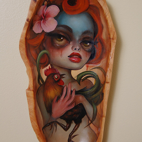 A surreal Tatiana Suarez painting on wood shows a woman with intense expression, vibrant red hair, elaborate makeup, holding a gallo. A pink flower decorates her hair while one side of her face is painted blue.