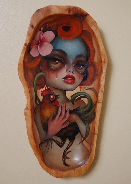 A surreal Tatiana Suarez painting on wood shows a woman with intense expression, vibrant red hair, elaborate makeup, holding a gallo. A pink flower decorates her hair while one side of her face is painted blue.