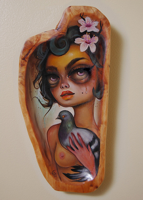 Tatiana Suarezs oil-on-wood painting depicts a stylized woman with dark eyes, surrounded by flowers and holding a palomita. The natural wood edge frames her soft, ethereal expression beautifully.