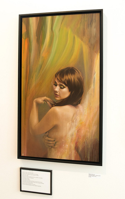 Nathan DeYoungs Sheltered Layers features a framed acrylic painting of a woman with short brown hair, gazing downward with crossed arms. The background shows soft shades of green, orange, and yellow. Displayed on a white wall with a plaque beneath it.