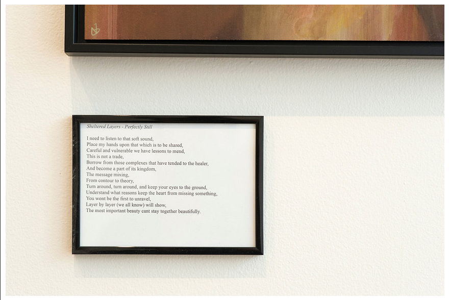 Nathan DeYoungs framed poem Silenced Layers - Perfectly Still, printed in black on white paper, is elegantly displayed in a thin black frame beside a vibrant acrylic painting, all set against a white wall.