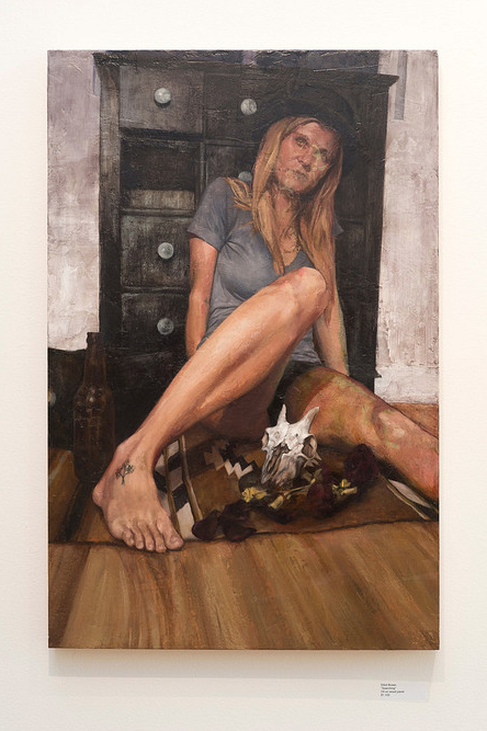 A painted portrait shows a woman on a wooden floor, leaning against a dark dresser. She wears a gray shirt and black hat. A bottle and animal skull with feathers rest by her tattooed foot, captured in detail suitable for an Elliot Brown gallery.