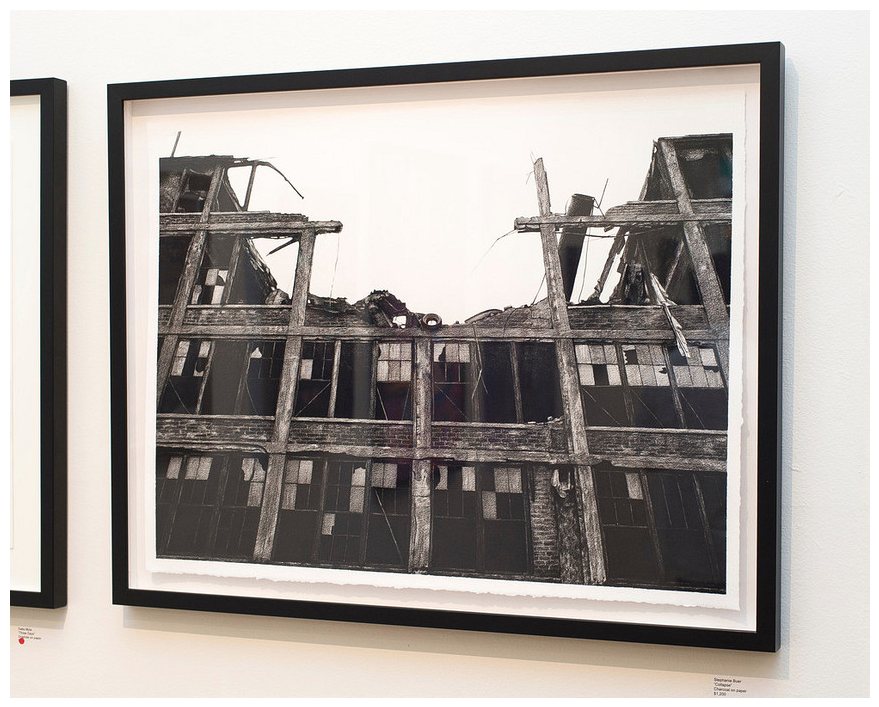 On a white wall hangs a black-and-white framed photograph of a dilapidated building with broken windows and crumbling structure, much like Stephanie Buers work, capturing an evocative sense of collapse and abandonment.
