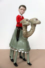 Doubleparlours hand-sculpted resin sculpture shows a boy in a red shirt and brown pants riding a unique two-headed, four-legged, camel-like creature with blank expressions. The boy also has a neutral expression against the plain background.