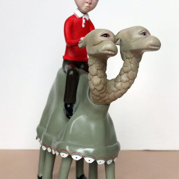 Doubleparlours hand-sculpted resin sculpture shows a boy in a red shirt and brown pants riding a unique two-headed, four-legged, camel-like creature with blank expressions. The boy also has a neutral expression against the plain background.