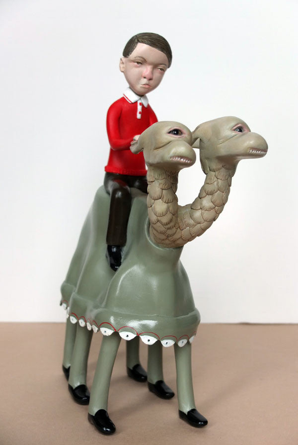 Doubleparlours hand-sculpted resin sculpture shows a boy in a red shirt and brown pants riding a unique two-headed, four-legged, camel-like creature with blank expressions. The boy also has a neutral expression against the plain background.