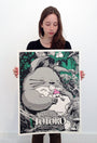 A young woman proudly holds a signed, numbered Joshua Budich screenprint of My Neighbor Totoro, featuring Totoro lounging in a forest with a small girl asleep on its belly amid lush greenery.