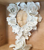 A serene face sculpture surrounded by white ceramic flowers, crafted in Stella Im Hultbergs distinctive style, is mounted on a wooden background. The intricate flowers form a heart-like pattern around the face.