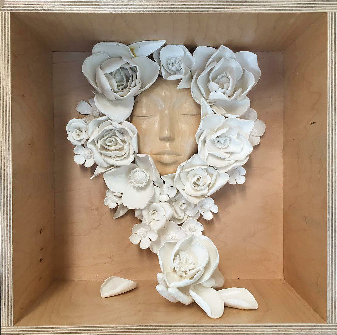A wooden box contains an art piece by Stella Im Hultberg, featuring a serene face surrounded by sculpted white flowers. The intricate floral arrangement frames the visage, with loose petals scattered like whispered secrets at the bottom.