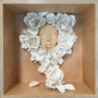 A wooden box contains an art piece by Stella Im Hultberg, featuring a serene face surrounded by sculpted white flowers. The intricate floral arrangement frames the visage, with loose petals scattered like whispered secrets at the bottom.