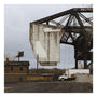 An urban industrial scene with a large concrete structure hanging from a steel bridge captures Crosshairs artistry in this limited edition of 65. Below, a white truck is parked by an empty lot, amid overcast skies and a small brick building.