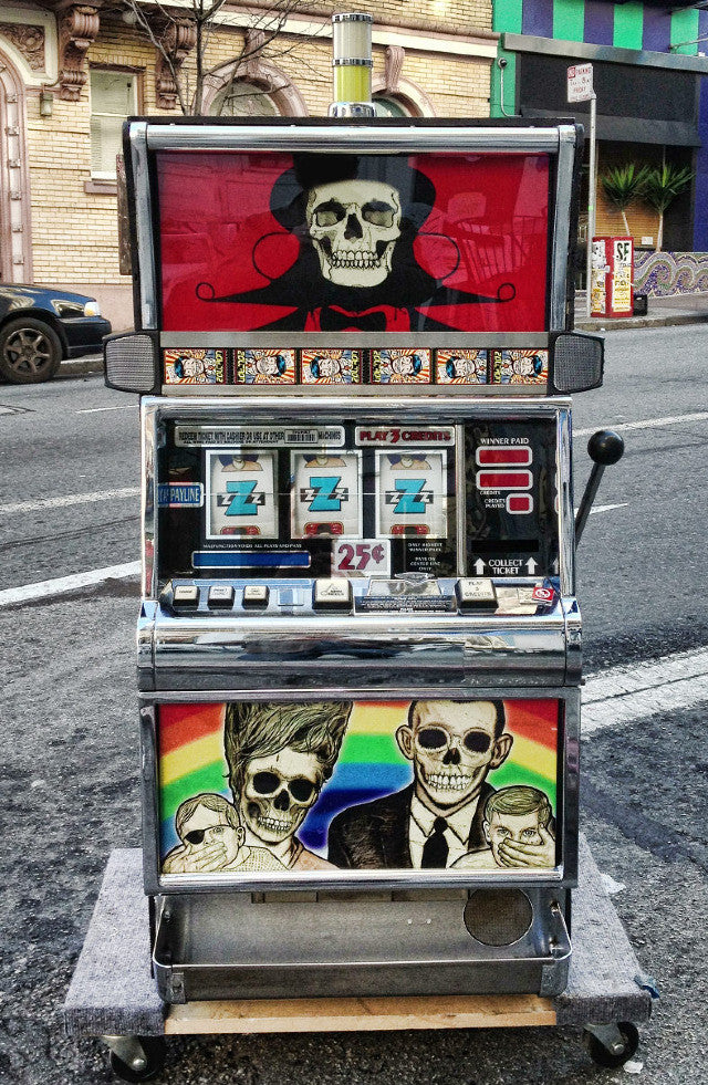 A vintage slot machine inspired by Zoltron features vibrant art of skulls in suits on a rainbow. A large skull with a mustache adorns the top, making it stand out on the sidewalk against a brick building backdrop.