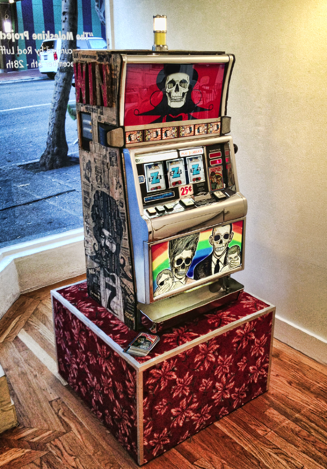 A Zoltron-crafted slot machine, featuring vibrant skull motifs, is displayed on a red patterned pedestal in an art gallery. Behind it, a window offers a glimpse of the street outside.